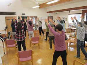 大勢の利用者さまが椅子から立ち上がってストレッチを行っている写真